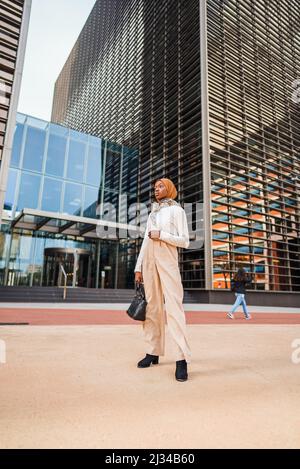 Ganzer Körper einer gut gekleideten afroamerikanischen Frau im muslimischen Kopftuch, die in der Nähe eines modernen Glasgebäudes in der Innenstadt steht und wegschaut Stockfoto