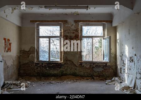 Blick in ein altes Zimmer mit großen Fenstern. Leerer Platz in einem verlassenen Gebäude. Ziegel kommen durch den Wandputz und die Farbe blättert ab. Stockfoto