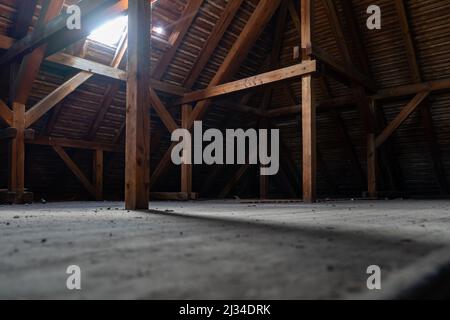 Alte staubige Dachboden eines Gebäudes. Leerer Raum, der durch ein Dachfenster beleuchtet wird. Holzkonstruktion des Dachs. Gruseliger Platz in einem großen Haus mit einer Horror-Atmosphäre Stockfoto