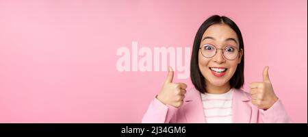 Super, Glückwunsch. Gesicht einer aufgeregten asiatischen Geschäftsfrau in einer Brille, lächelnd zufrieden, mit Daumen nach oben, stehend über rosa Hintergrund Stockfoto