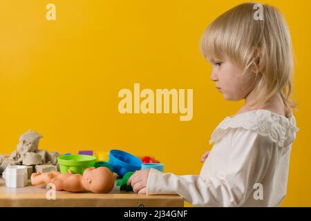 Ein schönes Mädchen in einem weißen Kleid steht neben einem Tisch mit kinetischem Sand, Plastikformen und einer Puppe. Gelber Hintergrund. Selektiver Fokus Stockfoto