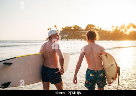 Vater mit Teenager-Sohn, der mit Surfbrettern am Sandstrand mit Palmen im Hintergrund, aufgehellt durch die Sonnenuntergangssonne, am Strand entlang läuft, haben sie ein Gespräch Stockfoto