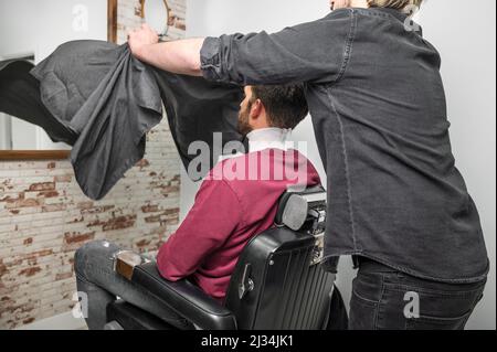 Hipster-Klient mit Umhang bedeckt wartet auf Frisur im Friseurladen. Hochwertige Fotos Stockfoto