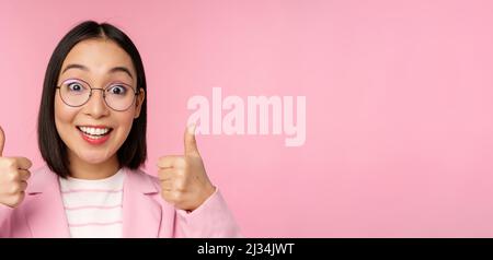 Super, Glückwunsch. Gesicht einer aufgeregten asiatischen Geschäftsfrau in einer Brille, lächelnd zufrieden, mit Daumen nach oben, stehend über rosa Hintergrund Stockfoto