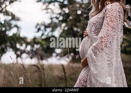 Eine Schwangere mit blonden langen Haaren im Wald trägt ein Kleid aus Boho-Spitze. Schönes Kleid und erwartete Frau im Freien in der Natur. Stockfoto