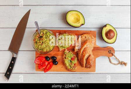 Flaches Foto, frisch zubereitete Guacamole in kleiner Glasschüssel, Brot, Tomaten, Oliven am Arbeitstisch und zwei Avocados am nächsten Schreibtisch aus weißem Holz Stockfoto