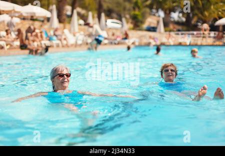 Zwei ältere Frauen mit grauem Haar, die im Hotelpool einen blauen Badeanzug tragen und auf dem Rücken schwimmen Stockfoto