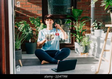 Junge Frau, die Yoga-Übungen macht und auf einer tibetischen Klangschale mit Schlägel in einem gemütlichen, modernen Loft-Zimmer voller heimeliger Pflanzen am sonnigen Morgen spielt. Stockfoto