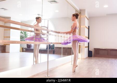 Die junge Ballerina engagierte sich im Tanzstudio. Eine hübsche Ballerina in einem violetten Tutu streckt sich in der Nähe der Theke, während sie auf ihren Zehen steht, sie ist Dämon Stockfoto
