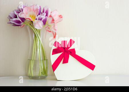 Ein Bouquet aus offenen rosa und violetten Tulpen, herzförmiger Geschenkbox und einer leeren Karte mit Platz für Text auf einem weißen Tisch Stockfoto