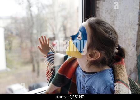 Ein kleines Mädchen mit einer ukrainischen Flagge auf ihrem Gesicht sitzt bedeckt mit einer Decke am Fenster. Kinder und der Krieg in der Ukraine Stockfoto