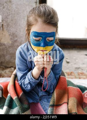 Ein kleines ukrainisches Patriotin mit der Flagge der Ukraine im Gesicht betet am Fenster. Kinder sind gegen den Krieg in der Ukraine. Stockfoto