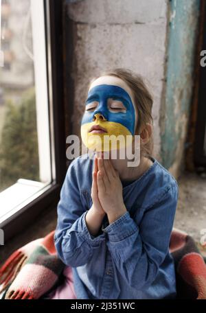 Ein kleines ukrainisches Patriotin mit der Flagge der Ukraine im Gesicht betet am Fenster. Kinder sind gegen den Krieg in der Ukraine. Stockfoto