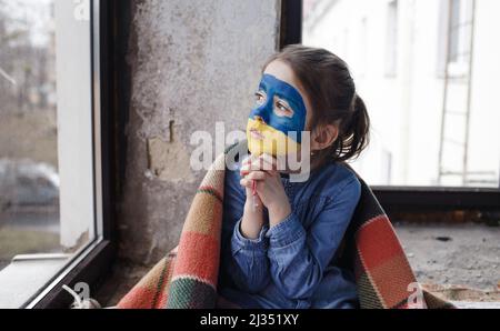 Ein kleines ukrainisches Patriotin mit der Flagge der Ukraine im Gesicht betet am Fenster. Kinder sind gegen den Krieg in der Ukraine. Stockfoto