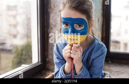 Ein kleines ukrainisches Patriotin mit der Flagge der Ukraine im Gesicht betet am Fenster. Kinder sind gegen den Krieg in der Ukraine. Stockfoto