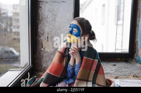 Ein kleines ukrainisches Patriotin mit der Flagge der Ukraine im Gesicht betet am Fenster. Kinder sind gegen den Krieg in der Ukraine. Stockfoto