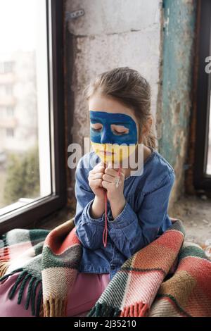 Ein kleines ukrainisches Patriotin mit der Flagge der Ukraine im Gesicht betet am Fenster. Kinder sind gegen den Krieg in der Ukraine. Stockfoto