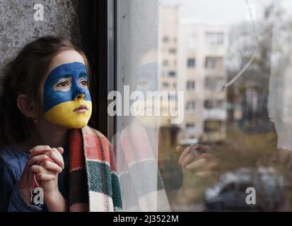 Ein kleines ukrainisches Patriotin mit der Flagge der Ukraine im Gesicht betet am Fenster. Kinder sind gegen den Krieg in der Ukraine. Stockfoto