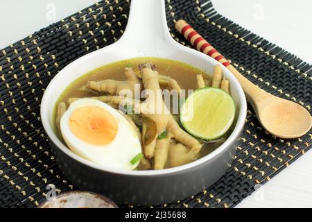 Indonesische traditionelle Küche namens Soto Ceker Ayam (Hühnersuppe mit Hühnerbein) auf grauer Keramikschale mit Sambal, gekochtem Ei und Limette Stockfoto