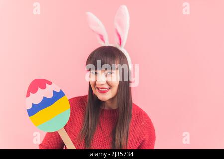 Ostern steht vor der Tür. Sympathische, langhaarige kaukasische Frau mit rotem Lippenstift, die vor der Kamera lächelt, während sie Hasenohren trägt und Ostereier aus Papier hält. Hochwertige Fotos Stockfoto