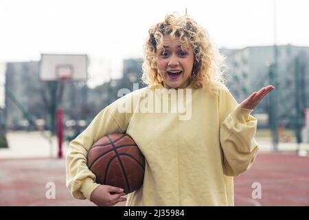 Verblüffte junge blonde Frau mit lockigen Haaren in einem hellgelben Pullover auf dem Sportplatz, die einen Basketballball hält und ihre andere Hand offen hält. Hochwertige Fotos Stockfoto