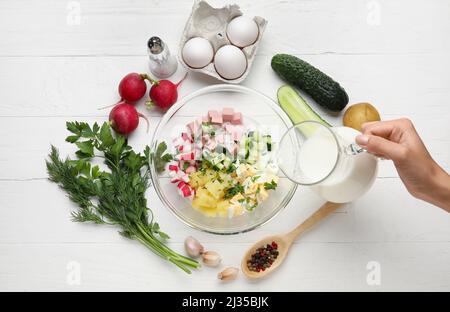 Frau Gießen Kefir in Schüssel mit Zutaten für Okroshka auf weißem Holz Hintergrund Stockfoto