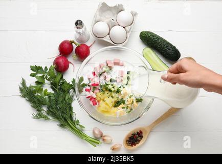 Frau Gießen Kefir in Schüssel mit Zutaten für Okroshka auf weißem Holz Hintergrund Stockfoto