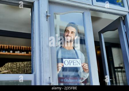 Ich kann es kaum erwarten, etwas großartiges zu servieren. Aufnahme eines hübschen jungen Mannes, der ein offenes Schild am Fenster seines Unternehmens aufgehängt hat. Stockfoto