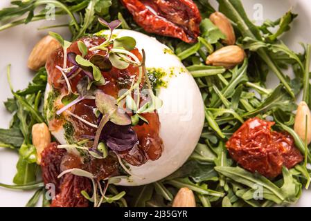 Typisch italienischer Burrata-Frischkäse-Salat mit Rucola, Sprossen, dehydrierten Tomaten mit Olivenöl, Pesto-Sauce und gebratenen geschälten Mandeln Stockfoto