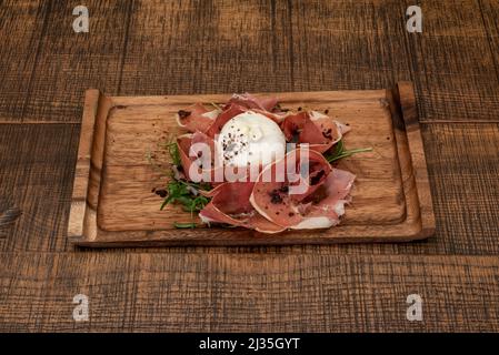 Typisch italienischer Salat aus frischem Burrata-Käse mit Rucola, gemahlenem schwarzen Trüffel und Scheiben serrano-Schinken auf einem Holzteller Stockfoto