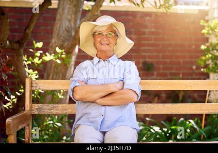 Im Garten zu sein bringt mir ein Lächeln ins Gesicht. Porträt einer glücklichen älteren Frau, die sich auf einer Bank im Garten entspannt. Stockfoto