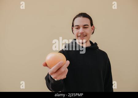 Junger, hübscher, großer, schlanker weißer Mann mit schwarzen Haaren, die Orangen im roten Hemd auf gelbem Hintergrund ausbalancieren Stockfoto