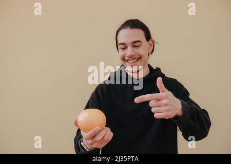 Junger, hübscher, großer, schlanker weißer Mann mit schwarzen Haaren, die Orangen im roten Hemd auf gelbem Hintergrund ausbalancieren Stockfoto