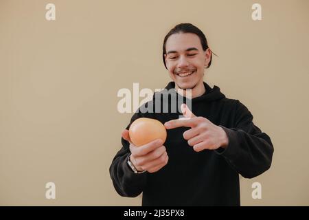 Junger, hübscher, großer, schlanker weißer Mann mit schwarzen Haaren, die Orangen im roten Hemd auf gelbem Hintergrund ausbalancieren Stockfoto