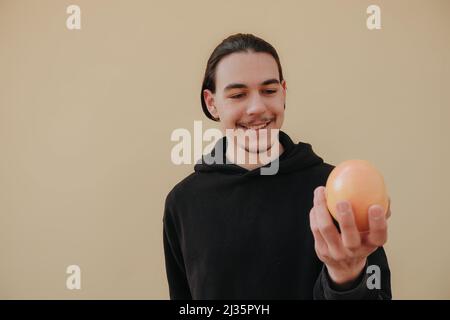 Junger, hübscher, großer, schlanker weißer Mann mit schwarzen Haaren, die Orangen im roten Hemd auf gelbem Hintergrund ausbalancieren Stockfoto
