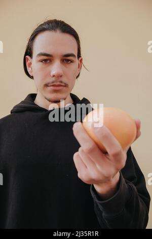 Junger, hübscher, großer, schlanker weißer Mann mit schwarzen Haaren, die Orangen im roten Hemd auf gelbem Hintergrund ausbalancieren Stockfoto