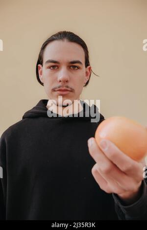 Junger, hübscher, großer, schlanker weißer Mann mit schwarzen Haaren, die Orangen im roten Hemd auf gelbem Hintergrund ausbalancieren Stockfoto