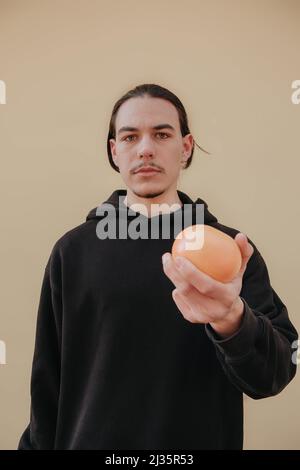 Junger, hübscher, großer, schlanker weißer Mann mit schwarzen Haaren, die Orangen im roten Hemd auf gelbem Hintergrund ausbalancieren Stockfoto