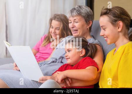 Großmutter liest ihren Enkeln ein Buch vor. Senioren und Mädchen auf der Couch lauschen Märchen Stockfoto