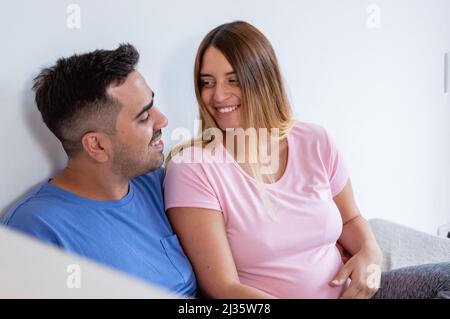 Der junge kaukasische Mann mit seiner Schwangeren Frau im Schlafanzug im Schlafzimmer sitzt auf dem Bett und schaut einander an und lächelt zusammen, kopiert Raum Stockfoto