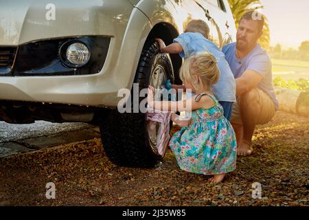 Sie sind immer bestrebt, Papa beim Waschen des Autos zu helfen. Aufnahme einer Familie, die ihr Auto gemeinsam wäscht. Stockfoto