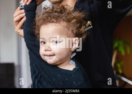 Kaukasisches Baby, das im Wohnzimmer steht und die Kamera im Profil betrachtet, während ihr Vater sie fertig kleidet. Stockfoto