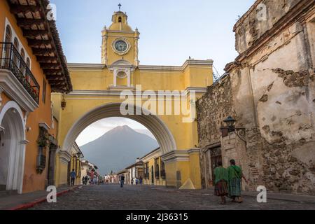 Menschen, die am frühen Morgen durch den Santa Catalina Arch gehen, um ins Stadtzentrum von Antigua Guatemala zu gelangen. Im Hintergrund ist der Vulkan Agua zu sehen Stockfoto