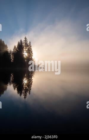 Ein nebliger Morgen über einem glatten See.die Morgensonne brennt durch die Bäume. Stockfoto