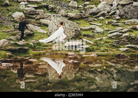 Frisch verheiratetes Paar, das auf einem felsigen Seeufer spazierengeht. Naturlandschaft. Eleganter, stilvoller Bräutigam und Braut in der Nähe des Flusses. Hochzeitspaar verliebt Stockfoto