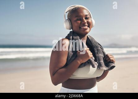 Ich arbeite an mir, für mich. Aufnahme einer sportlichen jungen Frau, die am Strand Kopfhörer und ein Handtuch um den Hals trägt. Stockfoto