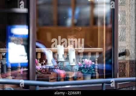 Riga, Lettland - 2. April 2022: Eine Frau an einem Tisch in einem Restaurant. Blick durch das Fenster. Straßensinnungen im Glas.