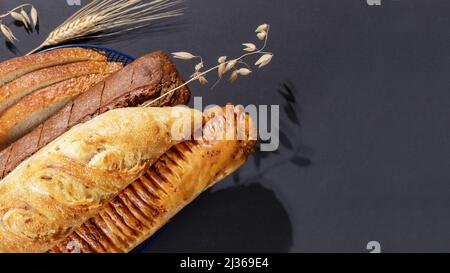 Hausgemachtes natürliches Brot. Frische Brotscheiben im blauen Korb mit Roggen- und Weizenohren auf schwarzem Hintergrund mit Kopierraum. Knusprige französische bagu Stockfoto