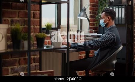 Buchhalter mit Gesichtsmaske Planung Marketing-Projekt auf Laptop bei der Firma Job. Büroangestellte erledigen Papierarbeiten mit Finanzstatistiken, um die Geschäftsentwicklung und das Wachstum zu fördern. Stativaufnahme Stockfoto