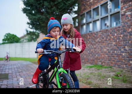 Ich werde Euch nicht fallen lassen. Eine kleine Aufnahme eines Mädchens, das ihrem Bruder beibringt, wie man draußen mit dem Fahrrad fährt. Stockfoto